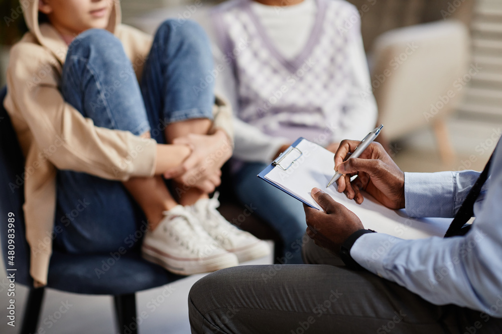 A therapist in a tie with a clipboard writing notes while a young girl sits with her parent, listening attentively.