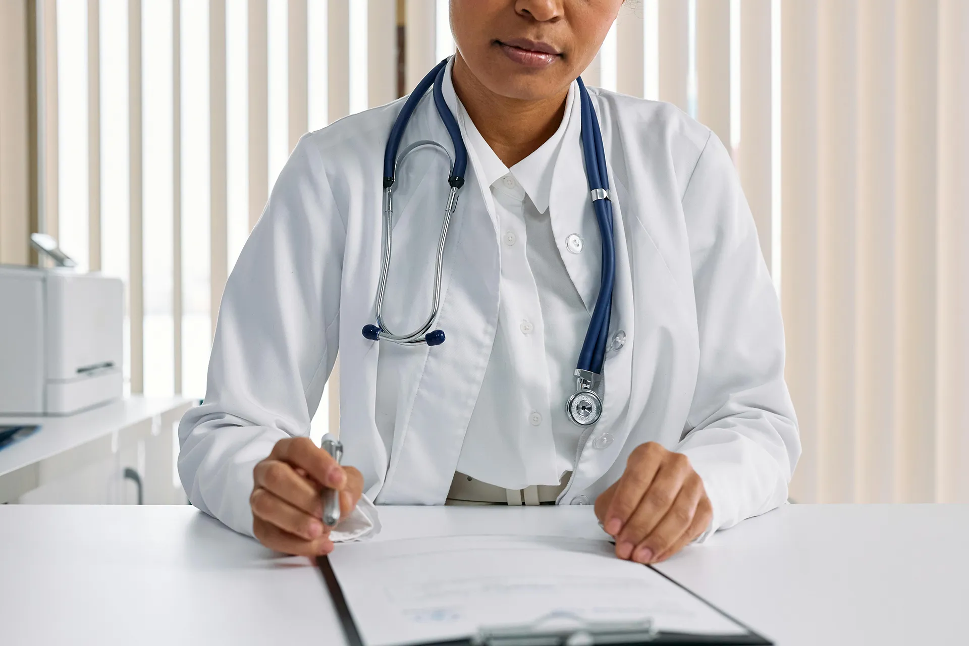 A doctor, wearing a white coat and stethoscope, is seated at a desk reviewing and signing a document.